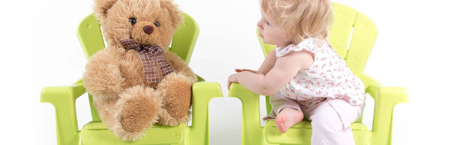 A child seated in a green chair looking at a teddy bear also seated in a separate green chair